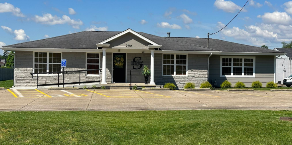 Sunset Elder Law's single-story office at 2806 New Hartford Road in Owensboro, Kentucky, has a gray stone front, white columns, and a large S monogram sign reading Sunset beside the black front door. A ramp with handrails and a parking space marked with a handicapped parking sign lead straight to the entrance, so clients can reach the office without climbing steps.