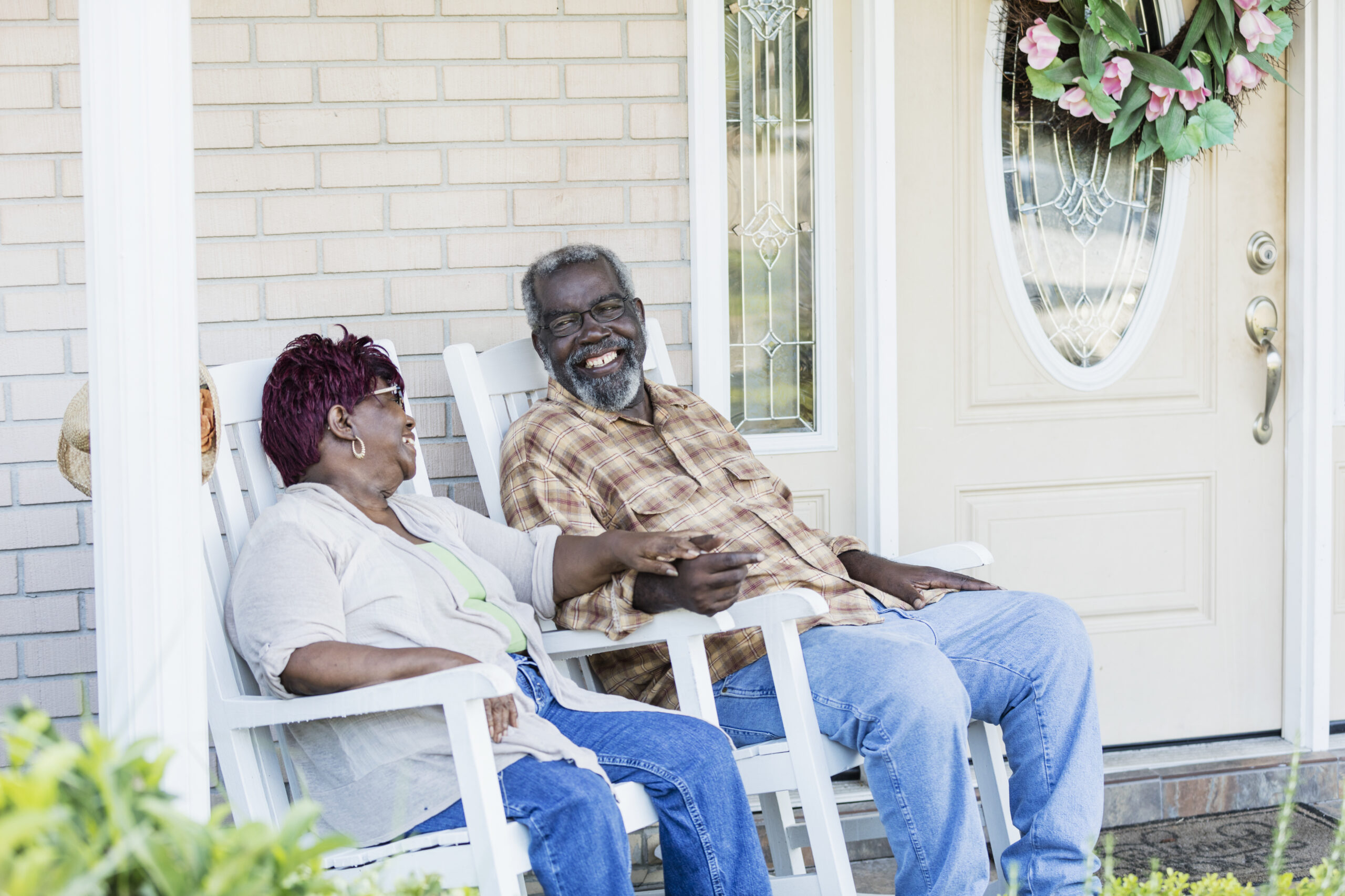 Holding hands in white rocking chairs, an older couple shares a laugh on the front porch of their brick home, a pink floral wreath on the door beside them. A Medicaid asset protection trust can help keep a home like theirs in the family if one of them later needs long-term care.