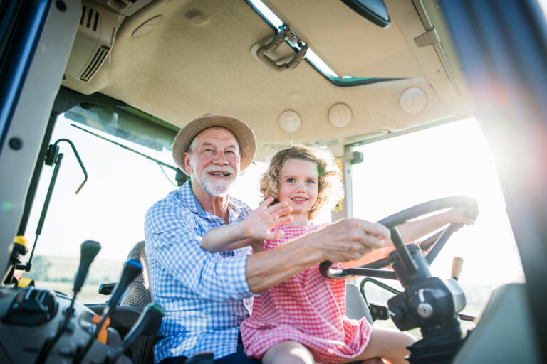 Seated on her grandfather's lap in a tractor cab, a curly-haired little girl grips the steering wheel and waves while he smiles beside her in a straw hat. An estate plan helps families pass the farm, and moments like this one, to the next generation.
