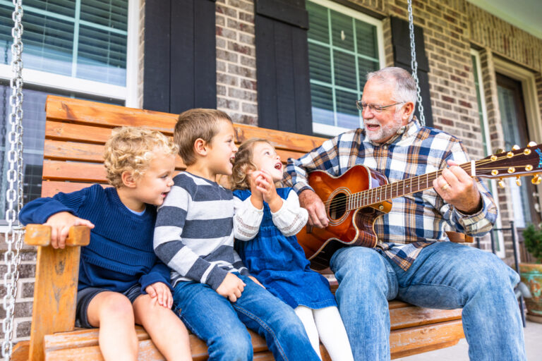Three young grandchildren sing and laugh on a wooden porch swing while their grandfather strums an acoustic guitar beside them. Medicaid planning helps older adults pay for long-term care while protecting the savings they built for their families.