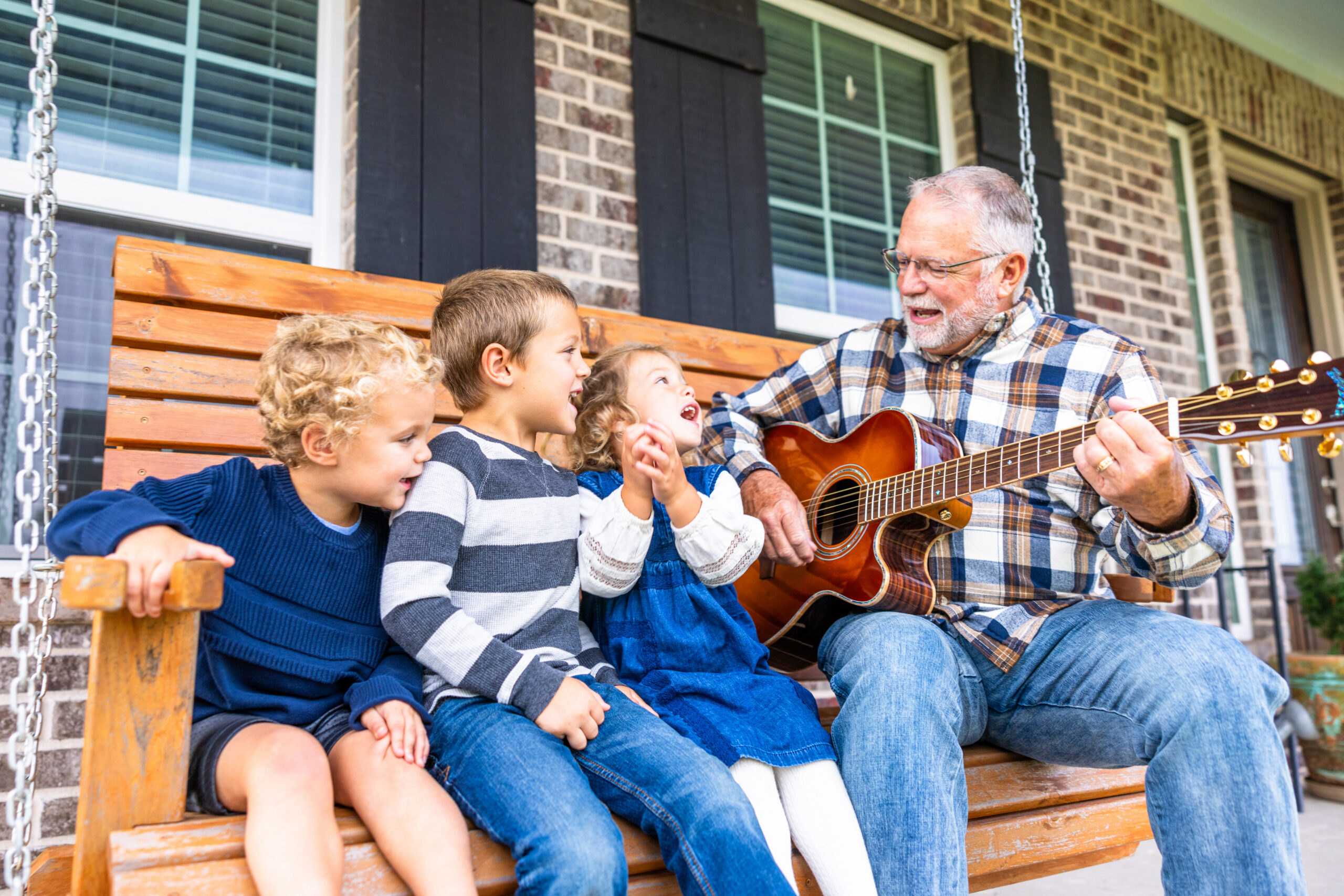 Three young grandchildren sing and laugh on a wooden porch swing while their grandfather strums an acoustic guitar beside them. Medicaid planning helps older adults pay for long-term care while protecting the savings they built for their families.