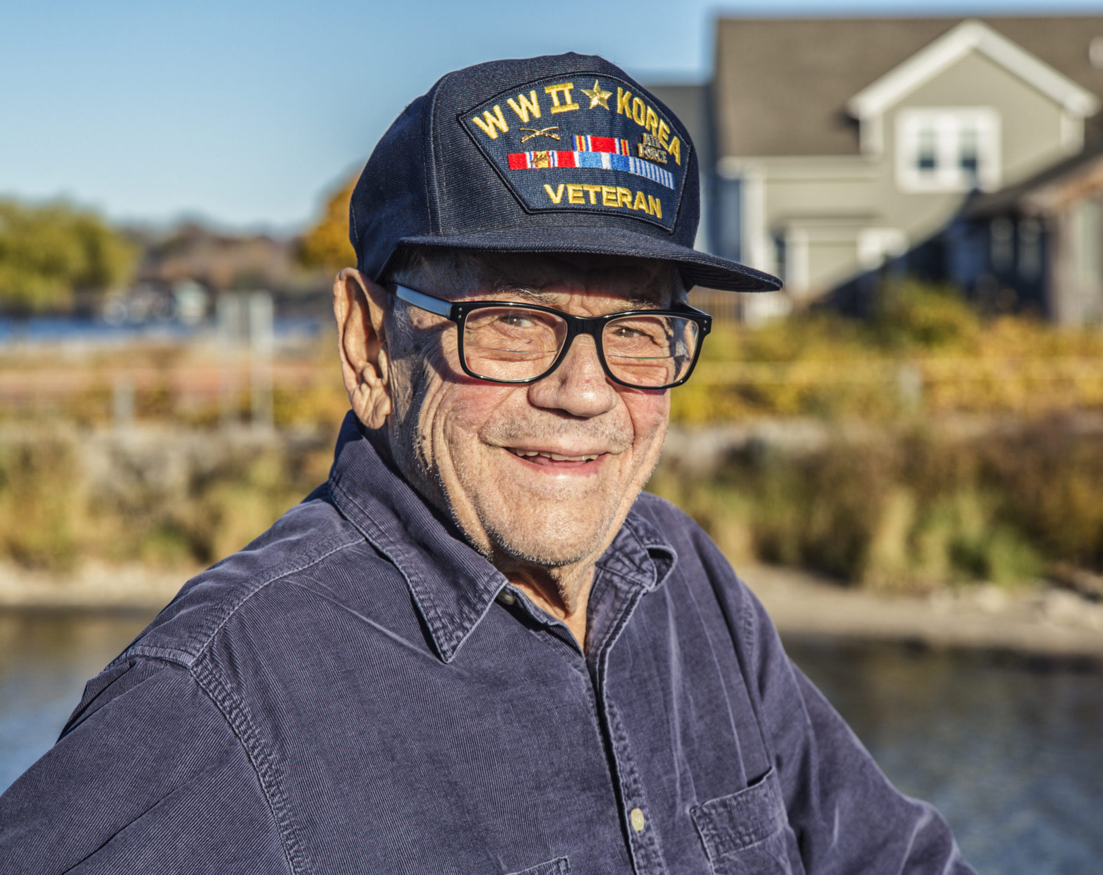 A smiling older veteran in dark-rimmed glasses wears a navy cap reading WWII Korea Veteran, embroidered with Air Force lettering and service ribbons. His wartime service is the kind that can help qualify a veteran and surviving spouse for VA benefits.